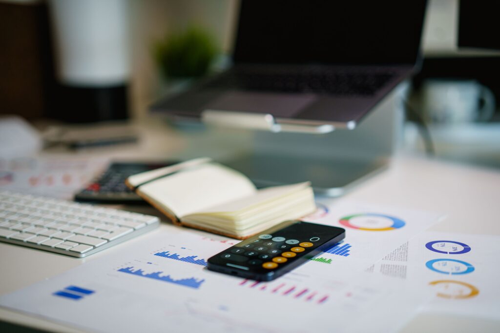 A close-up of an office desk with an open notebook, a keyboard, printed financial charts, and a smartphone displaying a calculator app, illustrating a PMO analyst focusing on financial planning and project salary bands.